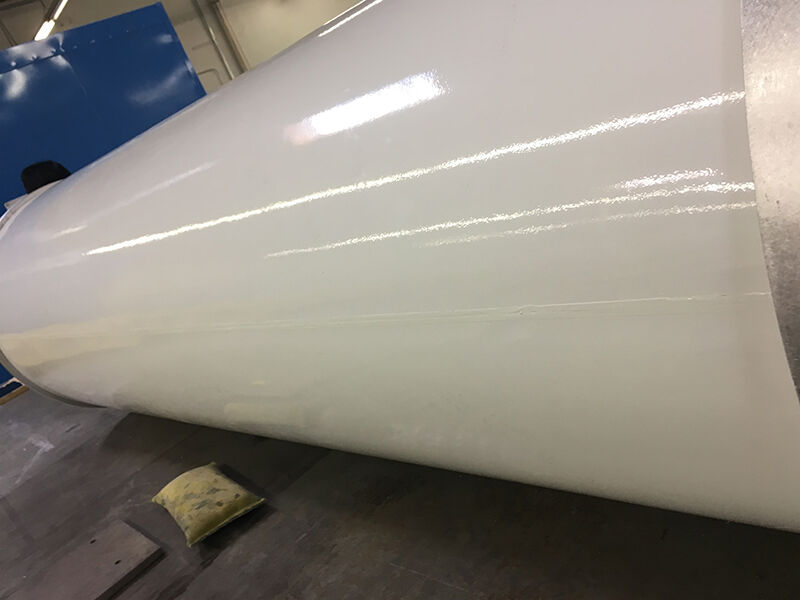 Image of the lamp post radome on its side, laying on the floor in a manufacturing space. It is a long white fiberglass tube with an aluminum metallic edge. There is a faint seam from edge to edge along the length of the radome. 
